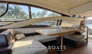 Spacious and airy interior of the 60’ Azimut Yacht ‘Tito’ with a large wrap-around couch, a wooden dining table, and panoramic windows offering views of the waterfront.