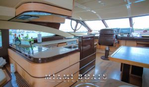 Interior of the 60’ Azimut Yacht ‘Tito’ showcasing the helm area with modern design features. The yacht’s bridge includes a captain’s chair, wooden paneling with a sleek finish, and a well-stocked bar with various bottles visible. Large windows provide a clear view outside. The “Miami Boats Rental” logo is prominently displayed.