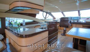 Interior of the 60’ Azimut Yacht ‘Tito’ showcasing the helm area with modern design features. The yacht’s bridge includes a captain’s chair, wooden paneling with a sleek finish, and a well-stocked bar with various bottles visible. Large windows provide a clear view outside. The “Miami Boats Rental” logo is prominently displayed.