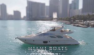 The 60’ Azimut Yacht ‘Wine Down’ glides through the sunlit turquoise waters of downtown Miami. With sleek curves, panoramic windows, and a spacious flybridge, the yacht radiates luxury and style. A guest relaxes on the upper deck as the city skyline and palm-lined waterfront create a striking backdrop for this elegant yachting experience.