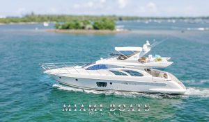 Side view of the 60’ Azimut Yacht ‘Wine Down’ cruising through the turquoise waters of Miami. The yacht features sleek, aerodynamic lines, expansive windows, and a spacious flybridge equipped with lounge seating and a sunshade. A rolled aqua mat rests on the upper deck, while a guest enjoys the rear lounge area. In the background, lush islands and scattered boats complete the tropical seascape.