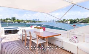 A birds-eye view of the 95' Dominator Super Yacht "Casual" in Miami, spotlighting the flybridge's well-equipped bar, wooden flooring, and plush sofas adorned with chic white and purple pillows.