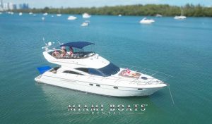 Luxurious 60 ft Viking Yacht for rental, named Princess, with people relaxing and sunbathing on the deck, cruising in the clear blue waters of Miami Beach, with a city skyline and other boats in the background. Miami Boats Rental logo is visible at the bottom of the image.