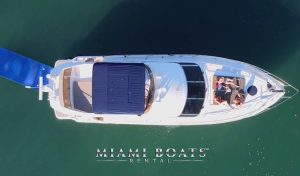 Aerial view of a luxurious 60-foot Viking Yacht named Princess, with people relaxing on the bow and under a canopy, anchored in the clear waters of Miami Beach. A blue water slide is extended from the yacht's stern. Miami Boats Rental logo is visible at the bottom of the image.