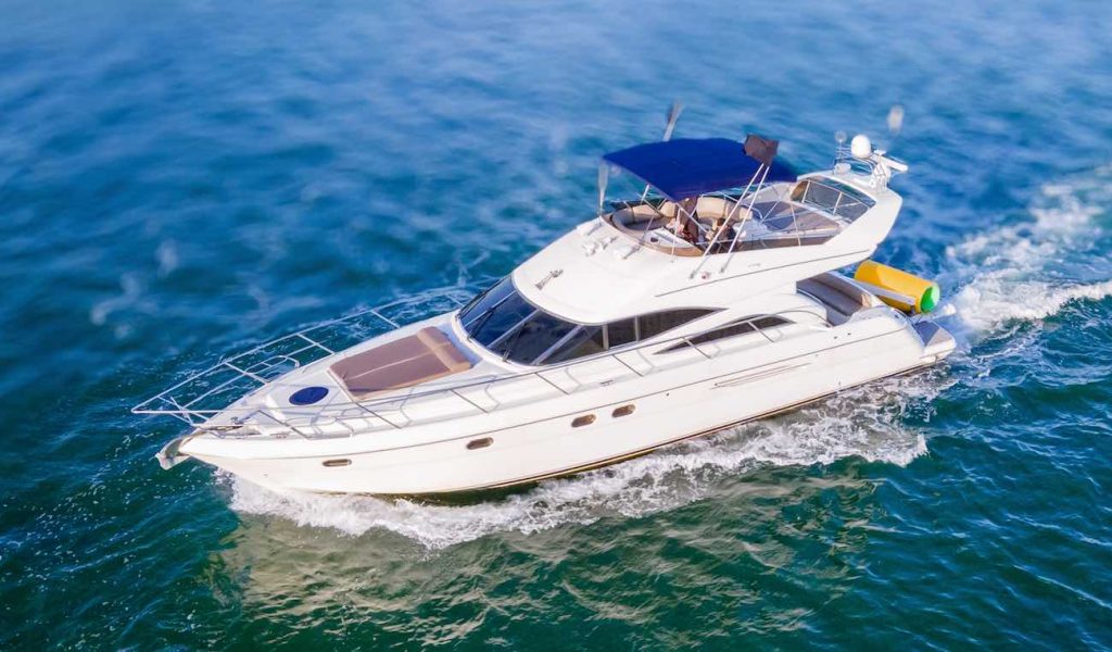 Aerial view of a 60' Viking Yacht Princess cruising through clear blue waters in Miami. The yacht has a sleek, white exterior with a blue canopy over the upper deck and spacious seating areas. The water is calm, creating a serene backdrop.