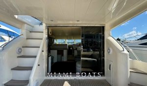 View of the entrance to the saloon of the 60' Viking Yacht for rental, named Princess, featuring a sleek, modern design with stairs on both sides leading to the upper deck. The entrance has sliding glass doors that open to the luxurious interior. The Miami Boats Rental logo is visible at the bottom of the image.