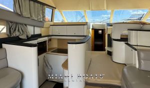 Interior of a luxurious 60' Viking Yacht Princess featuring a modern, stylish design. The image shows a spacious living area with plush seating, a compact kitchen with glossy white cabinets and countertops, and large windows allowing ample natural light. The "Miami Boats Rental" logo is visible at the bottom of the image.