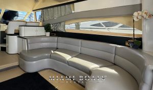 Interior of a 60' Viking Yacht Princess showing a sleek, modern design. The image features a curved, gray leather sofa, large windows with drawn curtains, and a glossy white cabinet with a potted plant on top. The "Miami Boats Rental" logo is at the bottom.
