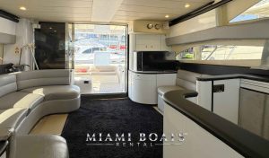 Interior of a 60' Viking Yacht Princess featuring a curved gray leather sofa, a TV on a glossy white cabinet, and a sliding glass door leading to the deck. The yacht's modern design includes large windows for ample natural light. The "Miami Boats Rental" logo is at the bottom.