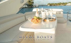 A luxurious yacht deck of Azimut Victoria setup on the water with "Miami Boats Rental" branding. The table features a fruit bowl with apples, oranges, and bananas, alongside a bottle of champagne, two wine glasses, and a tray, all set against the backdrop of a serene waterfront with lush greenery and docked boats in the distance.