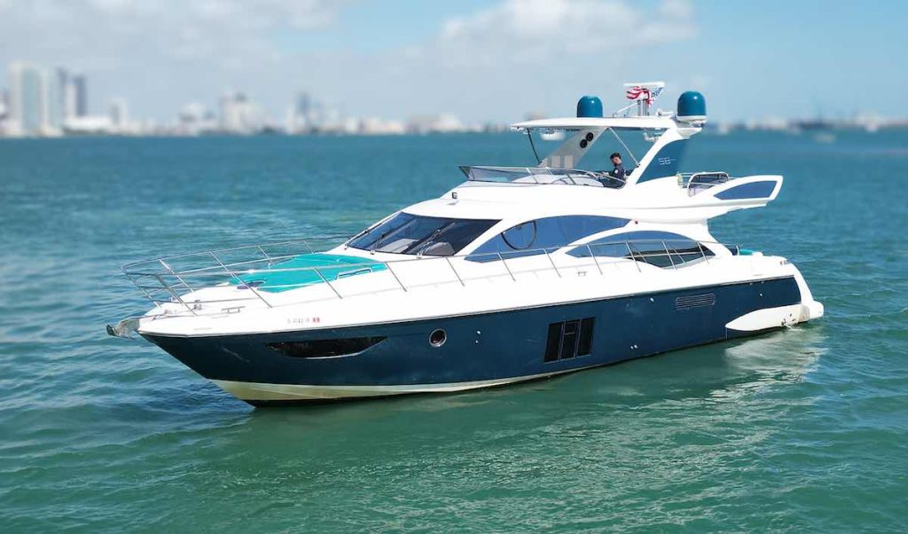 This image showcases the Luxury Yacht Azimut 'TITO' 60 luxury yacht, as it cruises on the serene waters off Miami's coast. The yacht features a striking dark blue hull with a white superstructure and teal accents that complement the vibrant Miami backdrop. The sleek, modern design of the yacht is highlighted by its streamlined windows and multiple decks, offering spacious areas for relaxation and viewing. The upper deck includes a helm station and additional seating, enhancing the outdoor boating experience. The photograph captures the essence of luxury yachting with Miami's skyline in the background, emphasizing a perfect blend of high-end leisure and the city’s dynamic atmosphere.