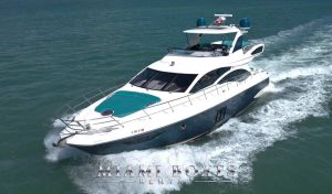 The 58ft Azimut yacht from Miami Boats Rental is captured cruising on the gentle waves of Miami's turquoise waters. The vessel's sleek white exterior with distinctive teal detailing, and an open bow area is complemented by the bright Miami sun. Passengers can be seen enjoying the ride from the flybridge, with the yacht leaving a foamy trail in its wake, signifying a blend of speed, comfort, and luxury.