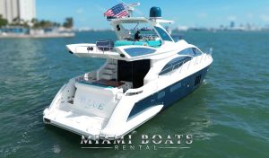 A pristine Azimut 60ft yacht from Miami Boats Rental is captured from the stern, showcasing its elegant white and navy blue design. The yacht is cruising in Biscayne Bay, with a man standing on the flybridge area against a backdrop of Miami's skyline. An American flag waves in the breeze above him, symbolizing the freedom and luxury of boating in Miami's vibrant coastal setting.