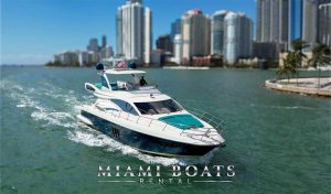 A white Azimut yacht for charter from Miami Boats Rental cruises through the turquoise waters of Biscayne Bay, with the Miami skyline in the background. The yacht features teal accents that complement the ocean's hue, and an American flag waves proudly on the top deck. The wake behind the yacht tells of its swift journey through the heart of Miami's waterfront, encapsulating the spirit of luxury and freedom at sea.