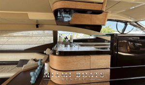 Modern interior of the 60’ Azimut Yacht TITO from Miami Boats Rental, showcasing a stylish bar and kitchen area with wood paneling, a curved countertop, and a selection of premium spirits. The space is accentuated by large windows and soft lighting, offering a refined and comfortable setting for entertaining at sea