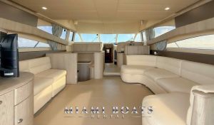 Luxury yacht rental in Miami – the elegant interior of a 60’ Viking yacht featuring a spacious salon with plush cream-colored leather seating, a flat-screen TV, modern wood cabinetry, and panoramic windows with ocean views. The galley kitchen and helm station provide a seamless blend of comfort and functionality. Ideal for private charters, corporate events, and luxury boat experiences. “Miami Boats Rental” branding is displayed at the bottom.