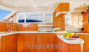 Interior view of the fully equipped galley on the 55’ Sea Ray Yacht - Island Itch, featuring glossy teak cabinetry, solid-surface countertops, a built-in sink, stovetop, and fruit bowl. Natural sunlight flows through large windows, enhancing the polished marine aesthetic.
