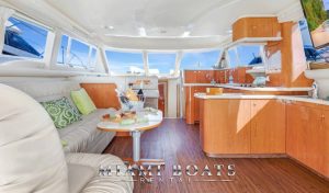 Wide interior shot of the Sea Ray 55’ Island Itch yacht showing an open-plan saloon with cream leather seating, a teak-paneled galley, and floor-to-ceiling windows. A charcuterie board and champagne setup complete the upscale ambiance.