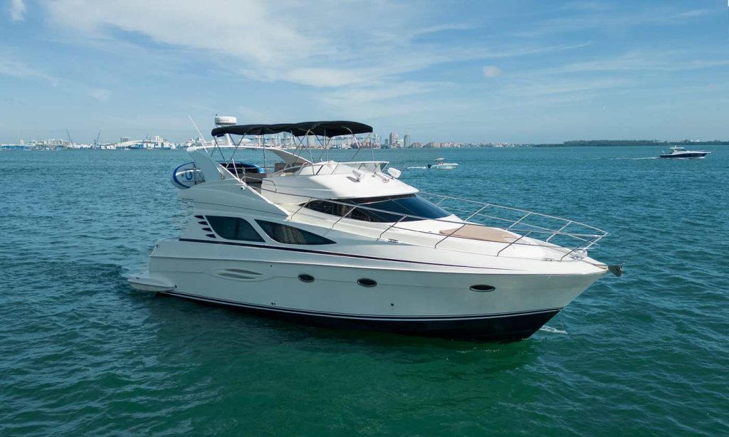 A 45-foot Silverton 'ESCAPE' yacht floats peacefully in the clear blue waters of Miami. This elegant vessel features a white hull with large windows and a spacious upper deck shaded by a black canvas bimini. In the background, the Miami skyline and other leisure boats provide a picturesque setting that embodies the essence of a tranquil sea escape.