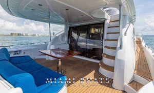 A view from the aft deck of a luxury yacht from Miami Boats Rental, showcasing an inviting outdoor lounge with plush blue cushions and a polished wooden table, ideal for socializing or relaxing. The open sliding glass doors lead to the yacht's elegant interior, with a glimpse of the Miami skyline in the background, highlighting an exquisite blend of indoor and outdoor luxury living.
