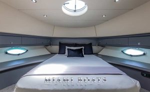 This image shows a meticulously arranged cabin inside the 80' Sunseeker Yacht 'SlipTease'. The cabin features a large bed with a custom gray and white bedspread, accented with a dark cushion for a touch of elegance. Above the bed, a skylight and side portholes provide natural light, enhancing the airy feel of the space. A flat-screen TV mounted on the opposite wall ensures entertainment is never too far away. The overall color scheme, a blend of gray tones with white accents, contributes to a modern, sophisticated ambiance, perfect for relaxation while cruising the waters near Miami.