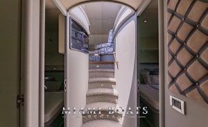 The image showcases a narrow corridor aboard the 80 ft Sunseeker Yacht 'SlipTease', presenting a stylish and modern interior. The corridor features a stairway lined with plush gray carpeting leading to an upper deck, accented with textured, taupe wall panels and minimalist chrome fixtures. An open door at the corridor's end reveals a glimpse of a cozy sleeping area, suggesting an inviting retreat below deck. The contemporary design and meticulous craftsmanship echo the luxury and comfort offered on this magnificent Miami yacht.