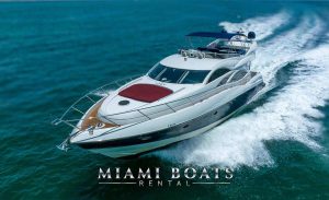 80-foot Sunseeker yacht named 'SlipTease' cruising at high speed through the open water, creating a wake behind it. The yacht features a sleek white and dark blue hull with a red sunbed on the bow and a spacious flybridge covered with a blue canopy. The logo of Miami Boats Rental is prominently displayed at the bottom of the image. 80' Sunseeker Yacht SlipTease Miami - Luxury Yacht Rental