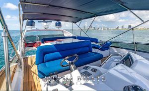 This image shows the flybridge area of an 80-foot Sunseeker Yacht named "Miami Boats Rental", cruising near Miami's coastline. The deck is equipped with plush seating upholstered in vibrant blue and white, providing a striking contrast against the rich wooden flooring. A large, shaded area offers respite from the sun, complete with modern navigational equipment and a clear view of Miami’s skyscraper-filled skyline in the distance. The setting is ideal for relaxing while enjoying the scenic ocean views and cityscape, highlighting the luxurious experience offered by Miami Boats Rental.