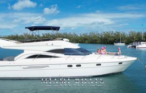 Luxurious 60' Viking Princess yacht cruising on clear blue waters in Miami, with a group of people relaxing on the deck, wearing pink hats. The yacht features sleek white design and spacious upper deck with blue canopy, epitomizing high-end boat rental experience.