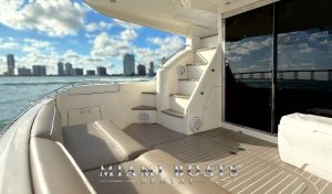 Luxury yacht rental in Miami – the spacious aft deck of a 60’ Viking Princess yacht featuring a comfortable seating area, elegant teak flooring, and a staircase leading to the flybridge. The stunning Miami skyline and a scenic bridge are visible in the background. Ideal for private yacht charters, special events, and luxury boat experiences. “Miami Boats Rental” branding is displayed at the bottom.