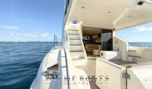 Luxury yacht rental in Miami – the spacious outdoor deck of a 60’ Viking yacht featuring sleek white railings, a comfortable seating area, and a staircase leading to the flybridge. The open sliding glass doors reveal a stylish interior with plush seating. Surrounded by the blue waters of Miami, this yacht is perfect for private charters, luxury boat rentals, and exclusive yacht experiences. “Miami Boats Rental” branding is displayed at the bottom.