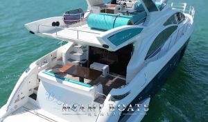 A rear-side aerial view of the 60’ Azimut Yacht from Miami Boats Rental, highlighting its spacious multi-level design. The aft deck features a cozy dining area with a wood table and teal-accented bench, while the flybridge above offers cushioned lounge seating and a rolled-up aqua mat for sunbathing. The yacht’s sleek curves and tinted windows enhance its modern elegance as it floats on the clear Miami waters.