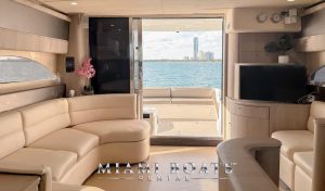 Spacious yacht salon with open deck – The stylish interior of a 60’ Viking yacht, featuring comfortable leather seating, a flat-screen TV, and modern wood cabinetry. Sliding glass doors lead to an outdoor deck with seating and ocean views, ideal for private yacht charters and luxury boat rentals in Miami. “Miami Boats Rental” branding is displayed at the bottom.